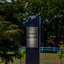 A dark metal sign for 'Welcome Studio Gallery' mounted on a black base, situated outdoors in a grassy area with trees and a white fence in the background.