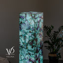 A tall rectangular Vilona lamp made of glowing green and purple marbled Fluorite stone, shown in a dim interior setting next to a potted plant.