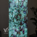 A tall rectangular Fluorite Vilona lamp made of polished natural stone with a marbled pattern of turquoise, mint green, purple, and brown, shown against a dark room background with a plant visible on the right.