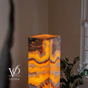A modern rectangular floor lamp made from glowing orange and yellow onyx with natural banding, set in a softly lit room next to a window and green plant. The brand logo 'VILONA' is visible in the bottom left corner.
