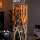 A tall rectangular Vilonà lamp made from glowing warm-toned onyx or agate with bold black, cream, and orange veining, shown in a dimly lit interior room. The Vilonà logo appears in the lower left corner.