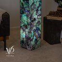 A tall rectangular Vilona floor lamp made of natural Fluorite with vibrant green, teal, and purple marbling, shown lit in a styled living room setting.