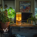 Decorative lamp with a stone-like pattern on a wooden table, surrounded by plants in a room setting.