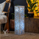 Decorative cylindrical lamp with a textured surface on a wooden table, surrounded by plants and a chair.