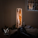 Angled lifestyle view of the illuminated White Ice onyx table lamp styled beside a window and lounge chair.
