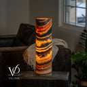 Illuminated Tangerine Onyx cylinder table lamp with sweeping orange, amber, and charcoal bands on a dark side table.