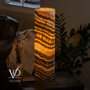 Illuminated Tangerine Onyx cylinder lamp on a dark side table beside a ceramic vase, book, and leather sofa.