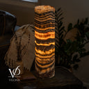 Glowing Tangerine Onyx cylinder lamp on a dark side table beside a ceramic vase, book, and leather sofa.