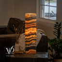 Illuminated Tangerine Onyx cylinder table lamp with a golden center, thin orange ribbons, and charcoal band above peach crystals.