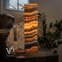 Glowing Tangerine Onyx cylinder lamp on a dark side table beside a ceramic vase, dried flowers, a book, and a leather sofa.