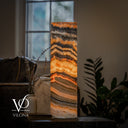 Glowing Tangerine Onyx cylinder lamp on a dark side table beside a ceramic vase, dried flowers, a book, and a leather sofa.