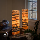 Tangerine Onyx cylinder table lamp pair illuminated beside a leather sofa, with amber, orange and charcoal bands.