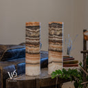Unlit Tangerine Onyx lamp pair with a pale crystalline formation interrupting the right lamp’s lower bands above its ivory base.