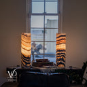 Wide front view of illuminated Tangerine Onyx lamps flanking books and a small vase beneath a window behind a leather sofa.