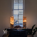 Wide room view of glowing Tangerine Onyx lamps beneath a tall window, with a leather sofa, stacked books, and green foliage.
