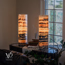 Angled view of illuminated Tangerine Onyx lamps beside a window, with the right lamp in the foreground and books and a cream throw between them.