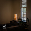 Aqua Onyx table lamp glowing beside books and dried stems near a tall window, with a leather sofa and greenery in the foreground.