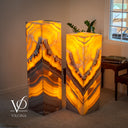 Two illuminated rectangular floor lamps made from polished onyx with natural banding in orange, yellow, cream, and gray, shown in a styled room setting with a window, side table, and plant. The brand name VILONA is visible as a watermark in the bottom left corner.