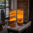 Two tall cylindrical table lamps made from layered orange, yellow, and brown agate-like stone, glowing warmly indoors on a stone-topped table in a rustic room. The brand name VILONA appears as a watermark in the lower left.