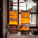 Two tall cylindrical Vases by Vilona made of natural agate with vibrant orange, yellow, and brown banding, displayed on a stone ledge in front of a window.