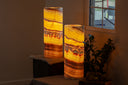 Two glowing Miami Sunset cylindrical lamp posts made from onyx stone, standing on a table in a dimly lit room beside a window and a plant. Made by Vilona Onyx.
